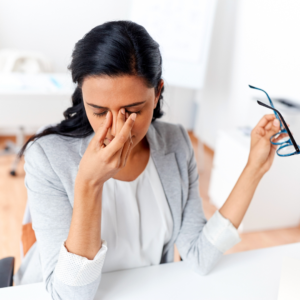 Woman in an office setting pinching the bridge of her nose and holding glasses, experiencing eye strain and fatigue as symptoms of iron deficiency in eyes