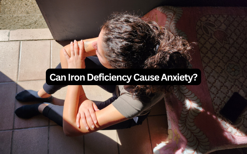 Woman sitting outdoors hugging her knees, with a text overlay reading "Can Iron Deficiency Cause Anxiety?"