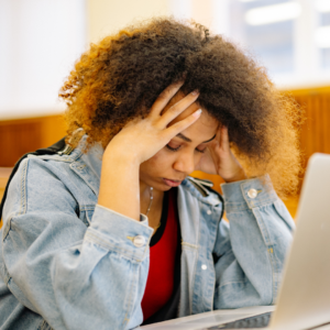 Woman in a denim jacket holding her head over a laptop, showing fatigue linked to causes of iron deficiency