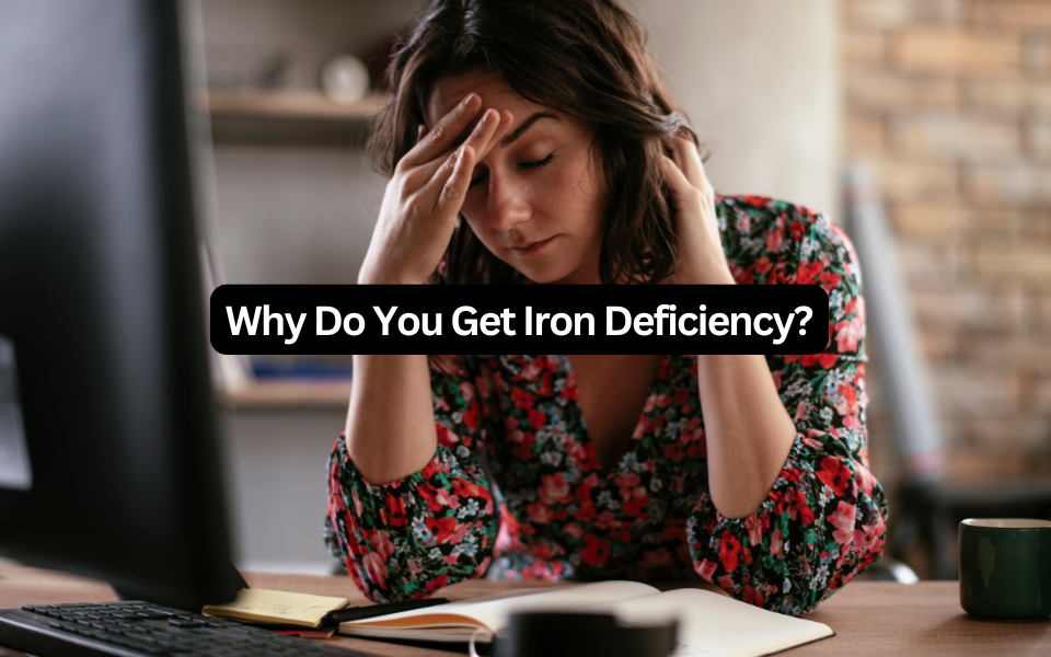 Why do you get iron deficiency? Tired woman in a floral blouse holding her head at a work desk